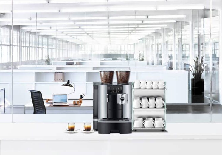 Kaffemaskin med bønnebeholdere og koppholder fylt med hvite kopper på en hvit benk i et lyst, moderne kontorlandskap.